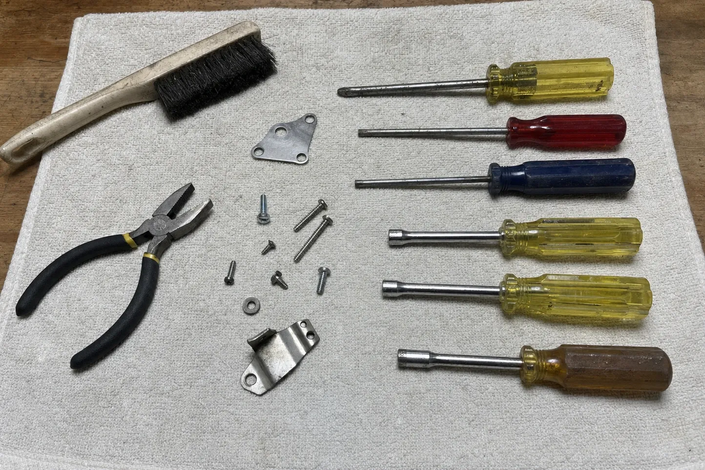 Screwdrivers, nut drivers, pliers and a wire brush laid out on canvas beside loose screws and two small stainless brackets